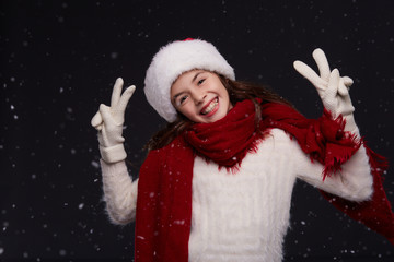 Portrait of young beautiful smiling girl in red Santa hat with white snowflakes on a dark background. Xmas fashion model with long curly hair. Winter holidays, Christmas, New Year concept