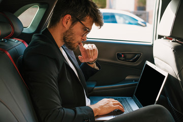 Attractive young man with a beard and glasses works with his laptop inside a car. Businessman.