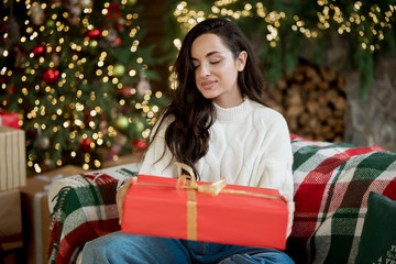 Young beautiful woman wearing warm sweater holding present sitting on the sofa in room decorated for celebrating the new year and christmas festive mood