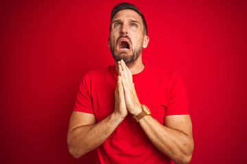 Young handsome man wearing casual t-shirt over red isolated background begging and praying with...