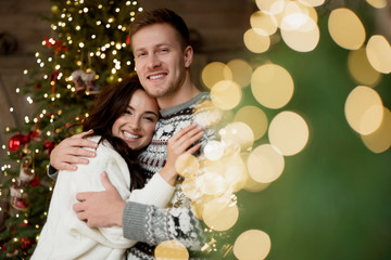 lovely couple husband hugging his beautiful wife standing near christmas tree both smiling in room decorated for celebrating new year festive mood love story