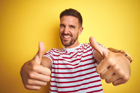 Young handsome man wearing casual red striped t-shirt over yellow isolated background approving doing positive gesture with hand, thumbs up smiling and happy for success. Winner gesture.