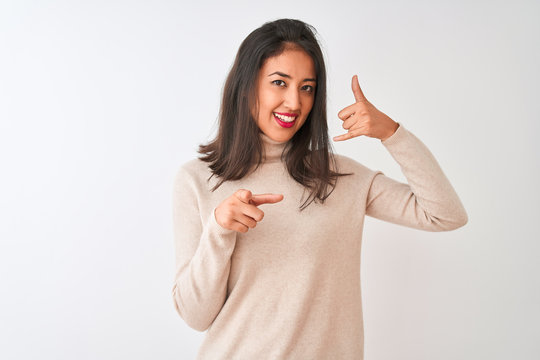 Beautiful chinese woman wearing turtleneck sweater standing over isolated white background smiling doing talking on the telephone gesture and pointing to you. Call me.