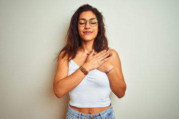 Young beautiful woman wearing t-shirt and glasses standing over isolated white background smiling with hands on chest with closed eyes and grateful gesture on face. Health concept.