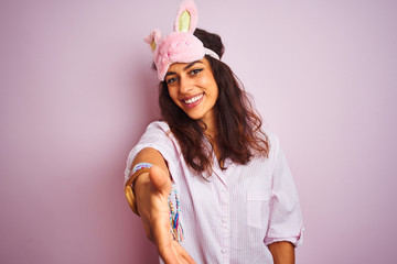 Obraz na płótnie Canvas Young beautiful woman wearing pajama and sleep mask over isolated pink background smiling friendly offering handshake as greeting and welcoming. Successful business.