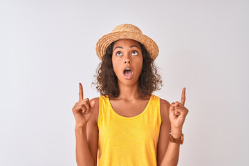 Young brazilian woman wearing yellow t-shirt and summer hat over isolated white background amazed and surprised looking up and pointing with fingers and raised arms.