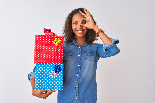 Young brazilian woman holding birthday gifts standing over isolated white background with happy face smiling doing ok sign with hand on eye looking through fingers