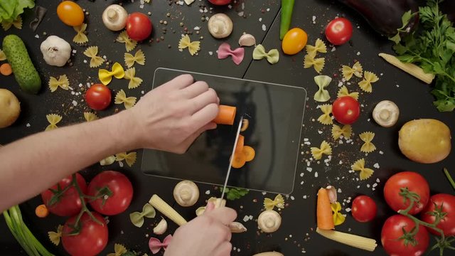 Overhead shot of crop Crazy cook cutting carrot on digital tablet, damaging the device and then stabbing knife at display on black table decorated, with vegetables and farfalle pasta