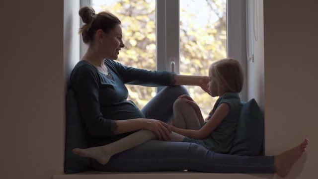 Pregnant woman caress daughter in turquoise clothes on windowsill