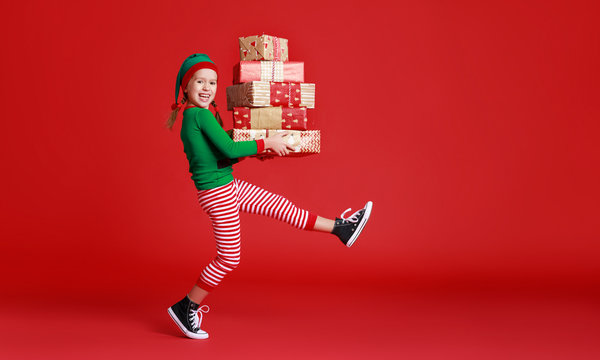 cheerful funny child in Christmas elf costume with gifts on   red background.