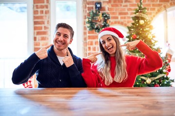 Young couple wearing santa claus hat sitting on chair and table around christmas tree at home smiling cheerful showing and pointing with fingers teeth and mouth. Dental health concept.