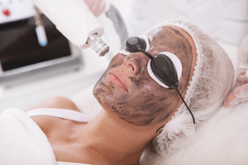 Close up of a mature woman wearing protective eyeglasses, getting carbon laser peel treatment by...