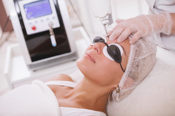 Close up of a beautiful woman in protective eyeglasses getting facial laser treatment by professional cosmetologist  - obrazy, fototapety, plakaty