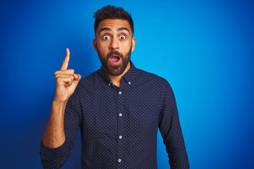 Young indian elegant man wearing shirt standing over isolated blue background pointing finger up with successful idea. Exited and happy. Number one.