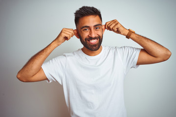 Fototapeta na wymiar Young indian man wearing t-shirt standing over isolated white background Smiling pulling ears with fingers, funny gesture. Audition problem