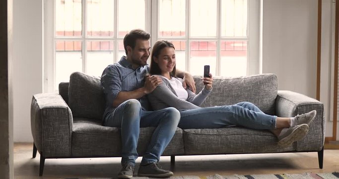 Relaxed millennial couple using phone taking selfie lounge on sofa