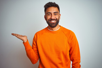 Young indian man wearing orange sweater over isolated white background smiling cheerful presenting and pointing with palm of hand looking at the camera.