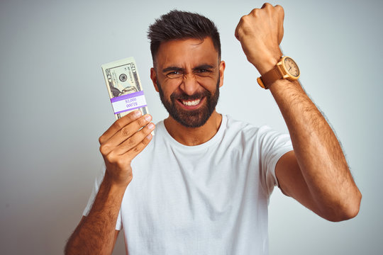 Young indian man holding dollars standing over isolated white background annoyed and frustrated shouting with anger, crazy and yelling with raised hand, anger concept