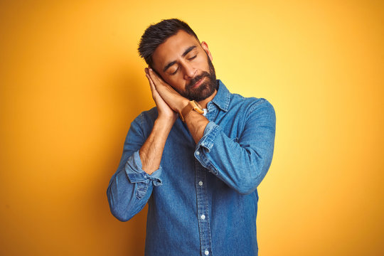 Young indian man wearing denim shirt standing over isolated yellow background sleeping tired dreaming and posing with hands together while smiling with closed eyes.