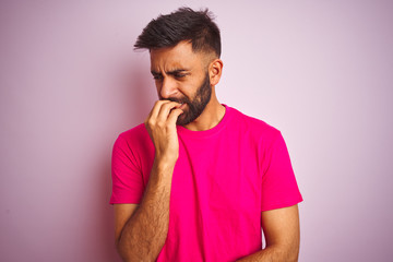 Young indian man wearing t-shirt standing over isolated pink background looking stressed and nervous with hands on mouth biting nails. Anxiety problem.