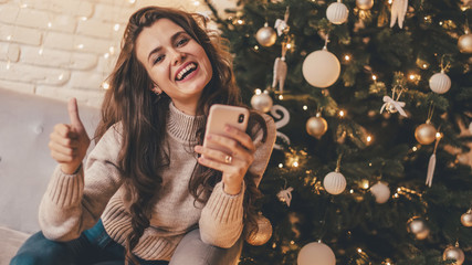 Woman with phone showing thumbs up sitting on the sofa near Christmas tree in the decorative...