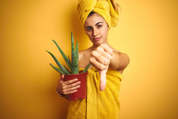 Young beautiful woman wearing a towel holding cactus pot over yellow isolated background with angry face, negative sign showing dislike with thumbs down, rejection concept