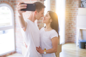 Young beautiful couple standing using smartphone to take a selfie at new home around cardboard boxes