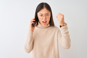 Young chinese woman talking on the smartphone over isolated white background angry and mad raising fist frustrated and furious while shouting with anger. Rage and aggressive concept.