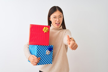 Young chinese woman holding birthday gifts over isolated white background approving doing positive gesture with hand, thumbs up smiling and happy for success. Winner gesture.