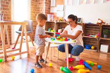 Beautiful teacher and toddler boy playing with construction blocks bulding tower at kindergarten