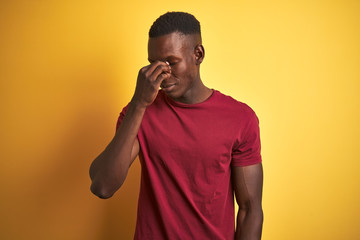 Young african american man wearing red t-shirt standing over isolated yellow background tired rubbing nose and eyes feeling fatigue and headache. Stress and frustration concept.