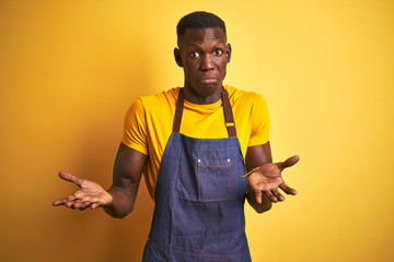 African american bartender man wearing apron standing over isolated yellow background clueless and...