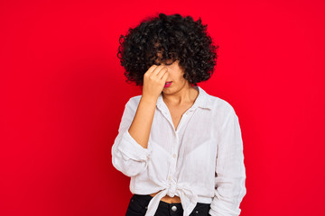 Young arab woman with curly hair wearing white casual shirt over isolated red background tired rubbing nose and eyes feeling fatigue and headache. Stress and frustration concept.