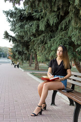Fototapeta na wymiar A young girl sits on a bench in a park and makes notes. Dressed in a free style. Business woman talking on the phone with customers. Time to relax and have some coffee