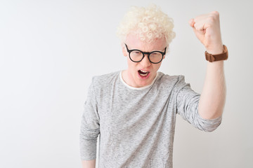 Young albino blond man wearing striped t-shirt and glasses over isolated white background angry and mad raising fist frustrated and furious while shouting with anger. Rage and aggressive concept.