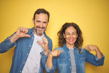 Beautiful middle age couple together wearing denim shirt over isolated yellow background looking confident with smile on face, pointing oneself with fingers proud and happy.
