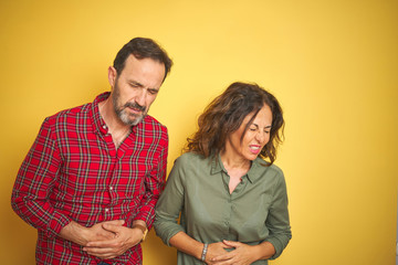 Beautiful middle age couple over isolated yellow background with hand on stomach because nausea, painful disease feeling unwell. Ache concept.