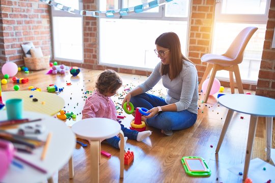 Young beautiful teacher and toddler sitting on the floor building pyramid using hoops at kindergarten