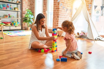 Beautiful teacher and blond toddler girl building tower using plastic blocks at kindergarten