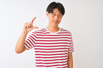 Young chinese man wearing glasses and striped t-shirt standing over isolated white background smiling and confident gesturing with hand doing small size sign with fingers looking and the camera.