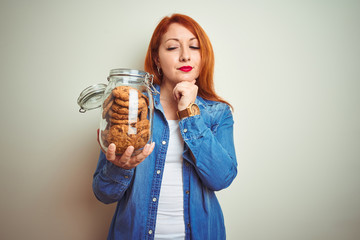 Young beautiful redhead woman holding jar of cookies over white isolated background serious face...