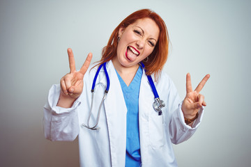 Young redhead doctor woman using stethoscope over white isolated background smiling with tongue out showing fingers of both hands doing victory sign. Number two.