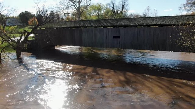 Aerial view floating down muddy flooded river, toward long wooden covered bridge