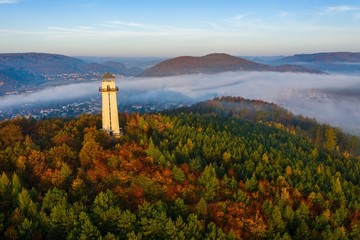White lighthouse in the middle of a tree forest near high grassy hills covered with fog