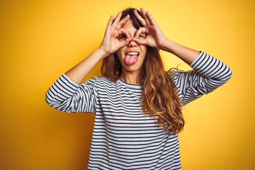 Young beautiful woman wearing stripes t-shirt standing over yelllow isolated background doing ok gesture like binoculars sticking tongue out, eyes looking through fingers. Crazy expression.