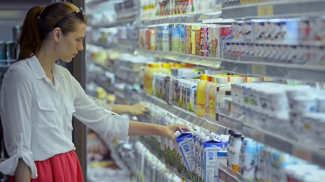 Young caucasian woman is choosing milk standing in grocery store. Beautiful female holding box in hand, watching content of dairy product and putting in basket. Side of housewife is on shopping walk