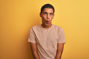 Young handsome arab man wearing striped t-shirt standing over isolated yellow background making fish face with lips, crazy and comical gesture. Funny expression.