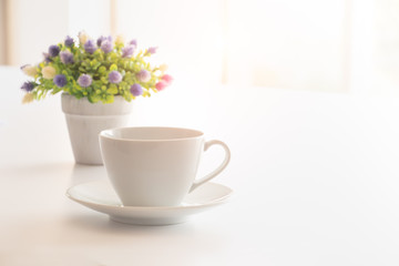 Fototapeta na wymiar A cup of coffee on white office desk with lovely vase of small flower with morning light flare. A good starting in the early morning to get ready for a day.Free copy space on right for text or design
