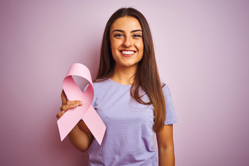 Young beautiful woman holding cancer ribbon standing over isolated pink background with a happy face standing and smiling with a confident smile showing teeth