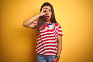 Young beautiful woman wearing striped t-shirt standing over isolated yellow background doing ok gesture shocked with surprised face, eye looking through fingers. Unbelieving expression.